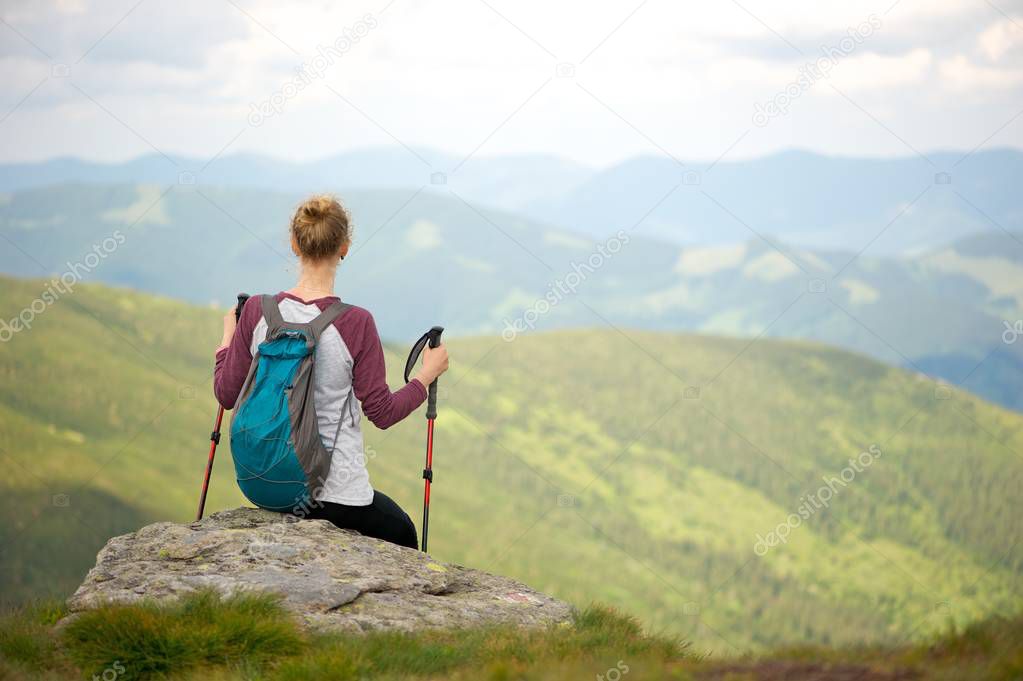 Short break on a hiking trip to enjoy the breathtaking view of the mountains. Young woman sitting on a rock, facing away from the camera; small backpack on her back and holding trekking sticks.