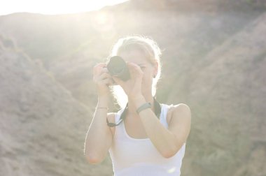 Bir yürüyüş gezisi fotoğraf çekmeye sarışın bir kadın. Beyaz kolsuz bluz, siyah kamera. Parlak güneş tepenin arkasından geliyor.