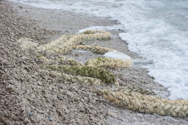 A closeup picture of an old role laying among stones and pebbles on a beach. Foamy sea water flowing towards it.