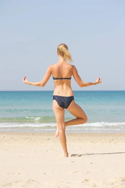Kamera arkası tek ayak üzerinde duran genç bir kadın; Bikini giymiş; Deniz arka planda.