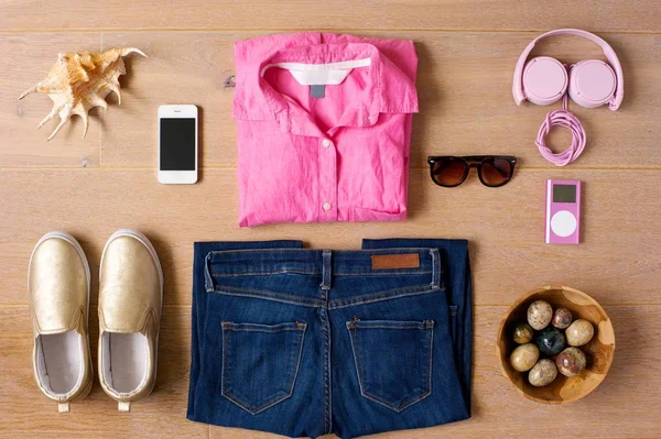 Casual kıyafet bir ahşap arka plan üzerinde ortaya koydu. Ayakkabı, kot pantolon, pembe gömlek, güneş gözlüğü, telefon, pembe müzik çalar ve kulaklık. Deniz kabuğu ve bir kase sidepieces olarak renkli taş. 