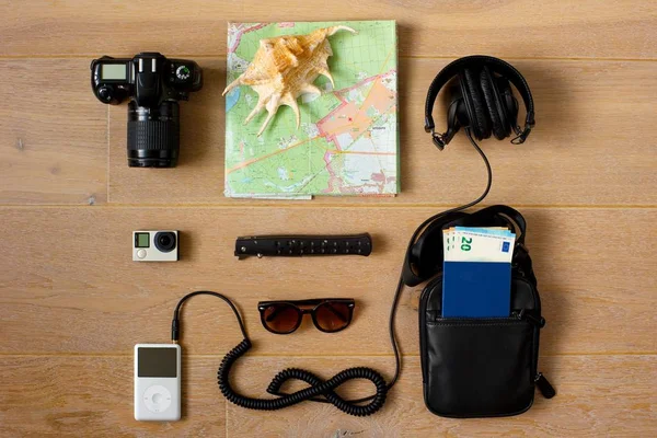 A traveler's kit. Photo and action cameras, music player with headphones, document bag with passport and money inside it, sunglasses, paper map and a sea shell. Top view. Light wooden background.