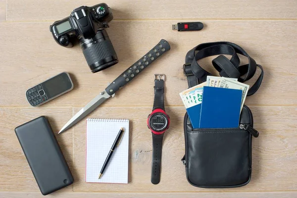 Ready-to-go spy kit.Photo camera, phone, powerbank, notepad with pen, knife, hand watch, USB memory stick, document bag with two passports and money in different currencies.Light wooden background.