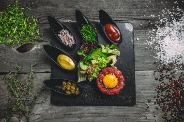 Siyah bir kare plaka üzerinde yumurta ile Dana gelen tartar. Soğan, ıspanak, marul, fesleğen, sos, hardal, Kapari, kekik, tuz ve baharat gri ahşap zemin üzerine