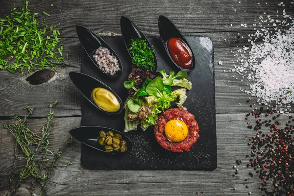 Siyah bir kare plaka üzerinde yumurta ile Dana gelen tartar. Soğan, ıspanak, marul, fesleğen, sos, hardal, Kapari, kekik, tuz ve baharat gri ahşap zemin üzerine