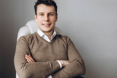 İyi bir ruh hali, çapraz kol ve gülümseyen bir gri duvara yakın oturan bir yakışıklı karizmatik adam portresi