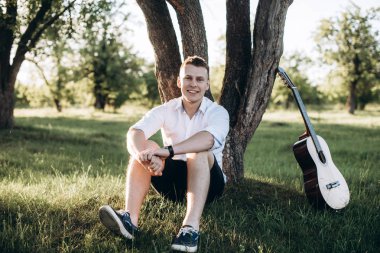 Gitar bir ağaç yakınındaki çimenlerin üzerinde oturan yakışıklı bir genç adam portresi ve gülümseyen 