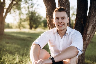 Sunset bulvarında bir ağaç yakınındaki çimenlerin üzerinde oturan yakışıklı bir genç adam portresi