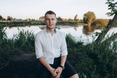 Günbatımı ve gülümsüyor bir göl oturan yakışıklı bir genç adam portresi