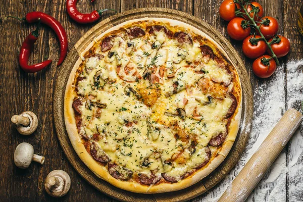 Delicious pizza with mushrooms, salami, cheese, tomatoes and pepper on a round wooden board on an old table. Top view