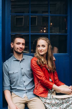 Oturan güzel mutlu çift duvar arka planda gülümseyen ve kameraya bakarak açık havada şık giysiler, mavi
