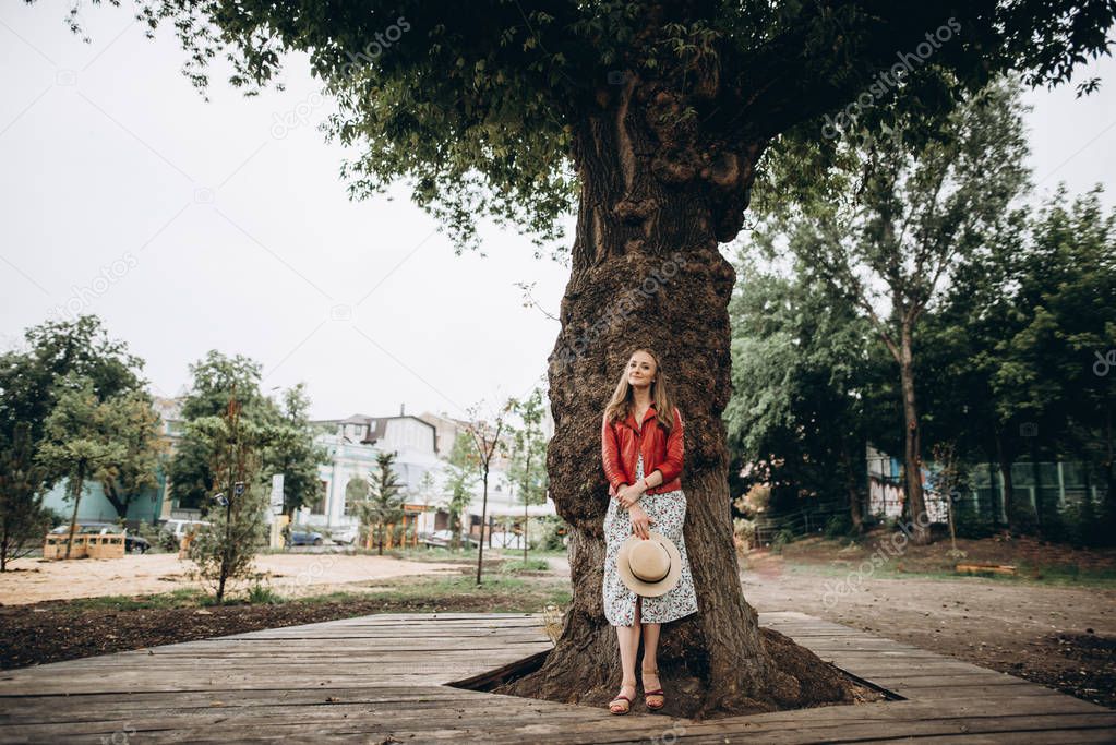 Beautiful young girl in a dress and a red jacket, with a hat in her hands, stands near a big tree and smiles