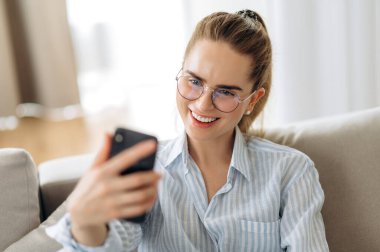 Telefon görüntülü arama. Genç ve harika bir kız rahat koltuğunda oturuyor ve arkadaşlarıyla telefon kullanarak video konferansında iletişim kuruyor.