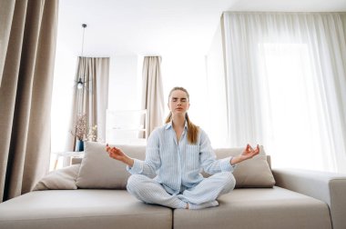 Evde yoga dersi var. Genç ve çekici bir kız, Lotus pozisyonunda meditasyon yaparken evdeki kanepede oturuyor.