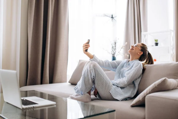 Phone video call. Smiling young caucasian girl in casual clothes and in a cheerful mood communicates via video calling by phone with friends while sitting at home on the couch