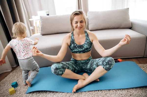 Sport and yoga at home. Mom practices yoga sitting in a lotus position on the floor, and her little adorable daughter plays nearby