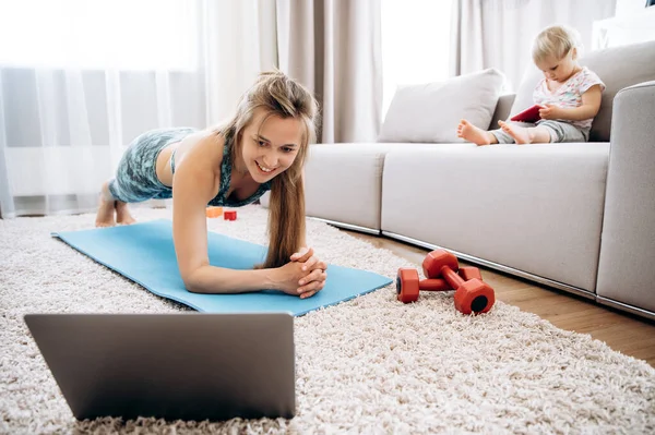 Sport at home. Young attractive mom doing sports at home and watching a video fitness lesson on a laptop, and her little baby girl sit on a couch