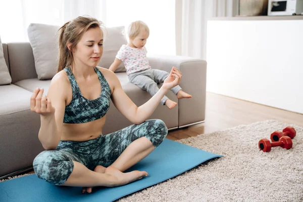 Sport and yoga at home. Mom practices yoga sitting in a lotus position on the floor, and her little adorable daughter plays nearby