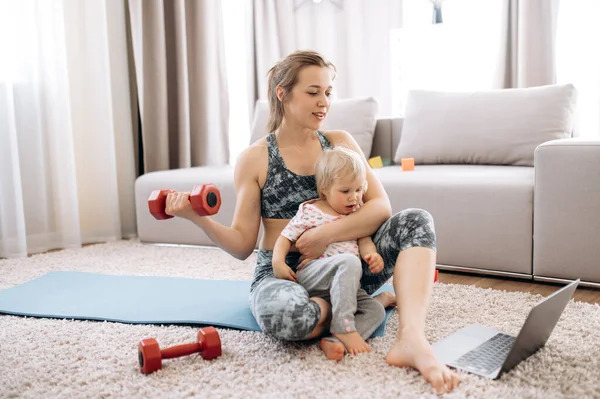 Home workout. Young attractive mom doing sports at home and watching a video fitness lesson on a laptop, with her little baby girl