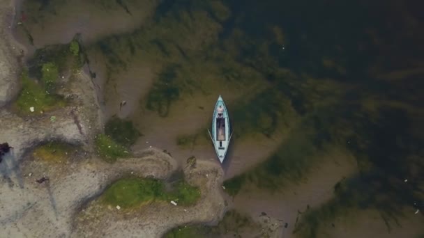 Un grand homme blanc se trouve dans un bateau bleu, sur la rive sablonneuse du Gange. Vue d'en haut, la caméra se lève verticalement. Varanasi, Inde .