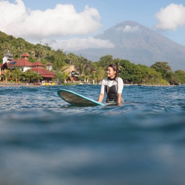 sportswoman sitting on surf board in ocean, coastline on background clipart
