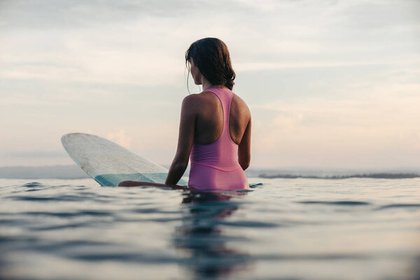 back view of girl sitting on surfboard in ocean at sunset