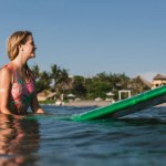 Zijaanzicht van de jonge vrouw in zwemmen pak rusten op surfen bord in oceaan met kustlijn op de achtergrond