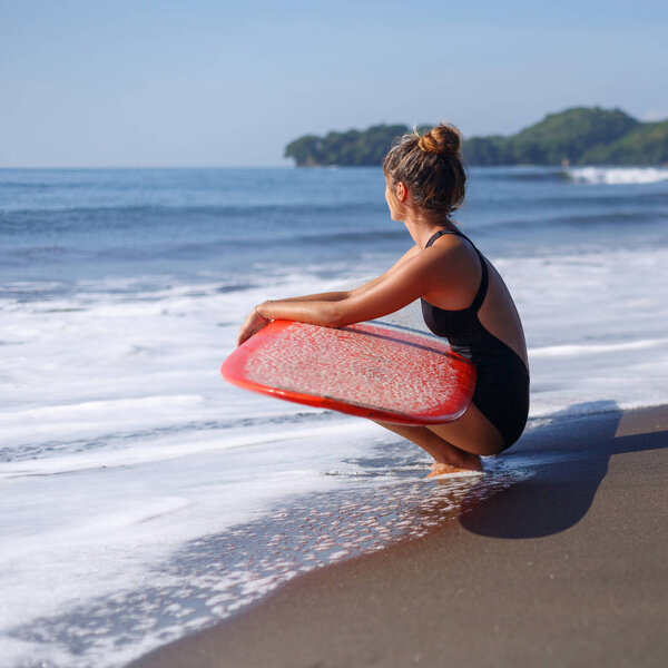 tanned surfer sitting with red surfboard on beach near ocean