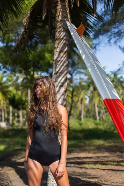 サーフボードのヤシの木のそばでポーズをとって髪の長い若い女性  — 無料ストックフォト