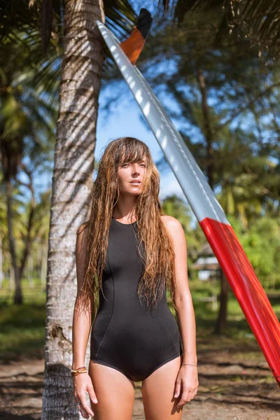 木の近くのサーフボードと水着でポーズ美しい日焼け少女 — ストック写真