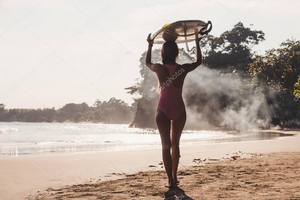 back view of sportswoman carrying surfing board on head while walking on coastline