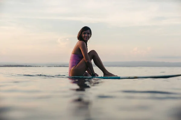 Belle fille assise sur la planche de surf dans l'eau dans l'océan au coucher du soleil — Photo de stock