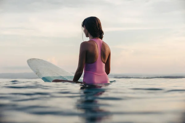 Vue arrière de la fille assise sur la planche de surf dans l'océan au coucher du soleil — Photo de stock