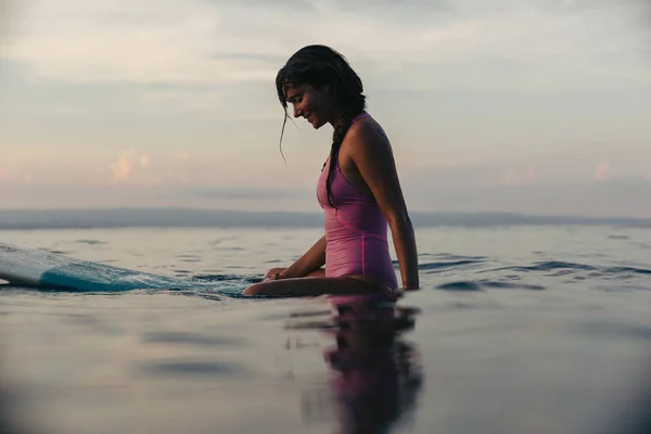 Menina desportiva sentado na prancha de surf na água no oceano ao pôr do sol — Fotografia de Stock