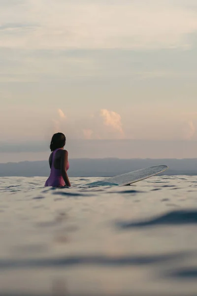 Vista traseira do surfista fêmea sentado na prancha de surf na água ao pôr do sol — Fotografia de Stock