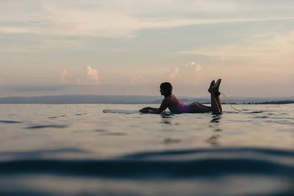 Silhueta de menina deitada na prancha de surf na água no oceano ao pôr do sol — Fotografia de Stock