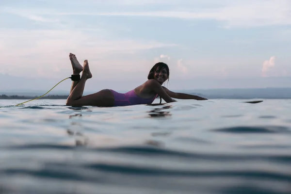 Улыбающаяся женщина, лежащая на доске для серфинга в воде в океане на закате — стоковое фото