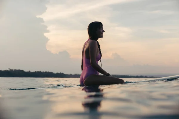 Surfista femenina feliz sentada en la tabla de surf en el agua al atardecer - foto de stock