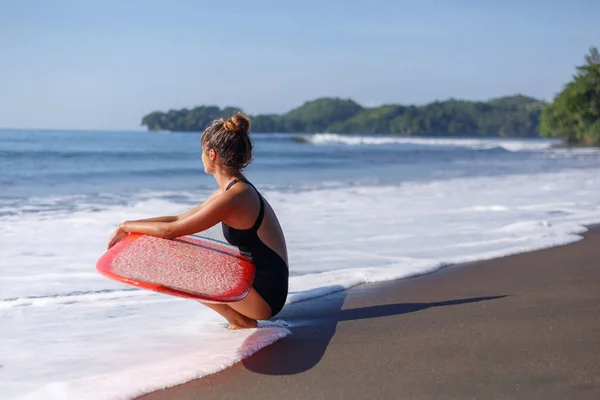 Jeune surfeur en maillot de bain assis avec planche de surf rouge près de la mer — Photo de stock