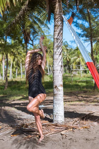 Attractive girl with long hair in black swimsuit posing with surfboard near palm tree — Stock Photo