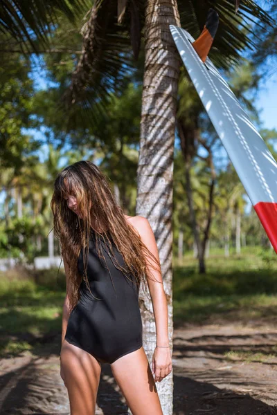 Femme surfeuse en maillot de bain noir posant avec planche de surf près de palmier — Photo de stock