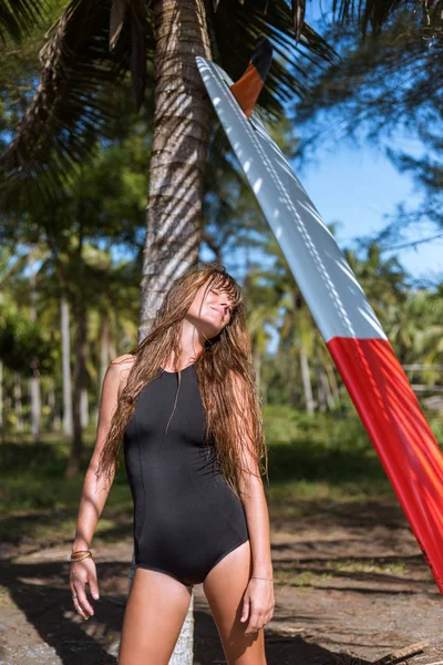 Smiling tanned girl posing in black swimsuit with surfboard near palm — Stock Photo