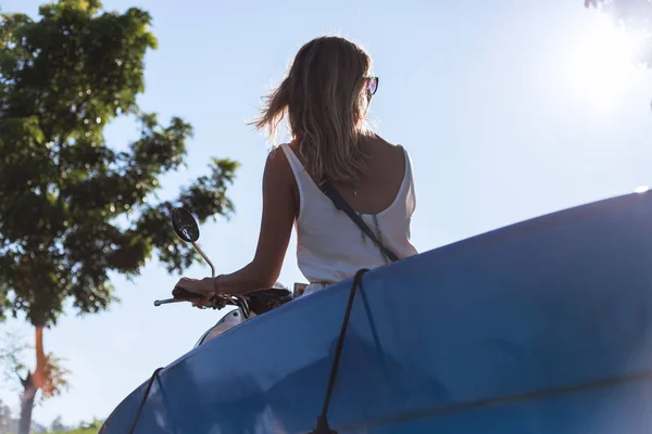 Back view of woman riding scooter with surfing board against blue sky — Stock Photo