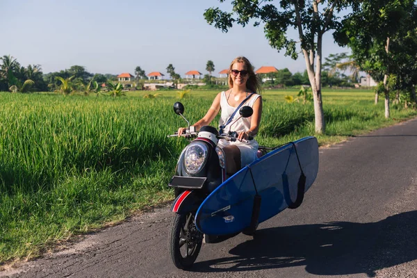Young smiling woman riding scooter with surfing board — Stock Photo