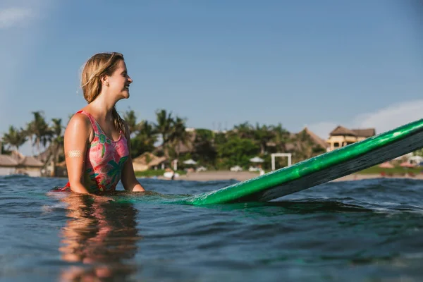 Vista laterale della giovane donna in costume da bagno appoggiata su tavola da surf in oceano con costa sullo sfondo — Foto stock