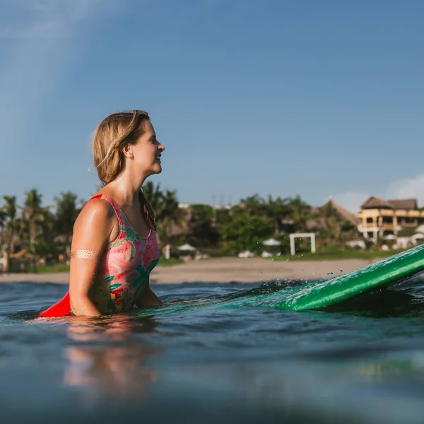 Vista lateral de la joven atractiva mujer en traje de baño descansando sobre tabla de surf en el océano con la costa en el fondo - foto de stock