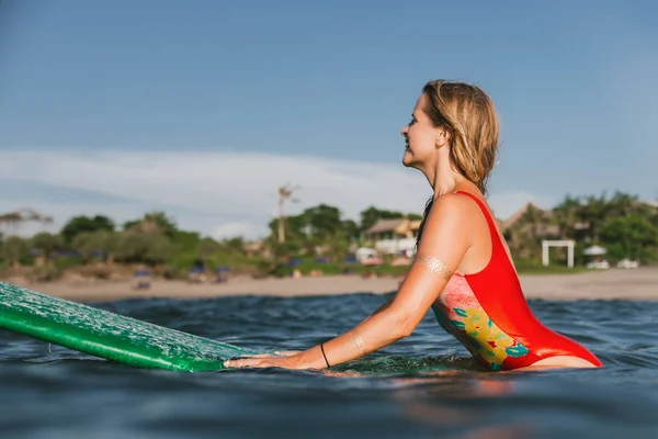 Side view of young attractive woman in swimming suit resting on surfing board in ocean with coastline on background — Stock Photo