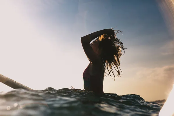 Silhouette de femme réajustant les cheveux tout en étant assis sur une planche de surf dans l'océan — Photo de stock