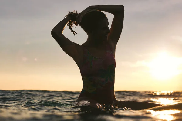Silhueta de mulher reajustando o cabelo enquanto sentado na prancha de surf no oceano ao pôr do sol — Fotografia de Stock