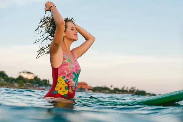 Молодая женщина в купальнике регулирует волосы, сидя на доске для серфинга в океане — стоковое фото